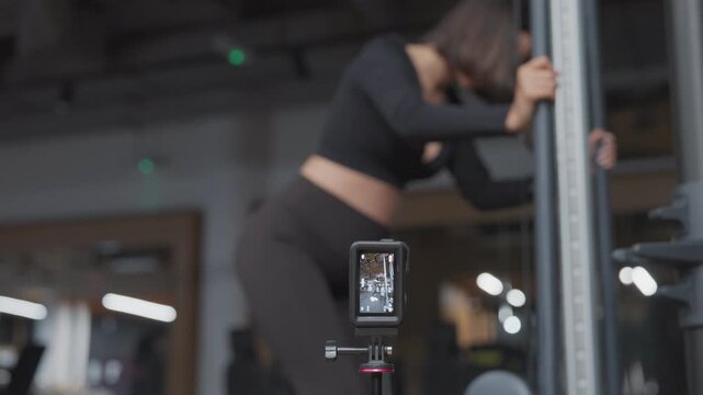 Action camera on a small tripod records a pregnant woman performing cable kickbacks in a gym in slow motion.