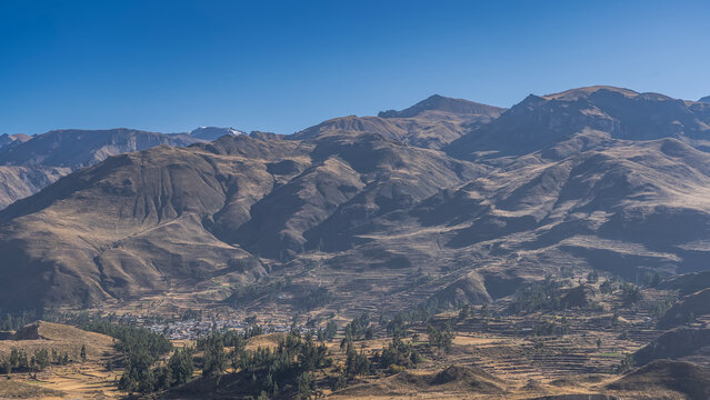 A beautiful highland landscape. The Andes mountain range against the backdrop of a blue sky. Agricultural terraces on the slopes. Rural buildings and green trees in the valley. Peru.