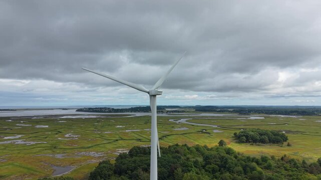 Single wind turbine spins above green coastal marsh and winding tidal creeks under heavy gray clouds captured from around with drone.