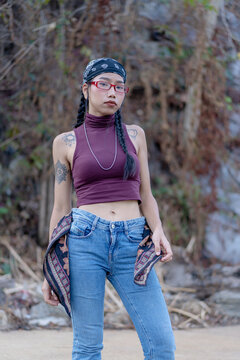 Trendy young Asian woman standing outdoors with a natural background, wearing a crop top and jeans with a stylish bandana, glasses and tattoos, posing in urban street style fashion.