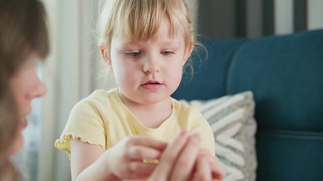 Young girl in yellow shirt sits on blue couch. She bites her nails nervously while looking down. Mother's hand holds her fingers gently. Girl shows signs of anxiety or boredom