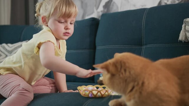Young girl gently feeds orange cat on sofa. Child looks focused, calm, and thoughtful. Cat eats with gentle interest, head lowered. Scene captures tender, quiet moment between child and pet