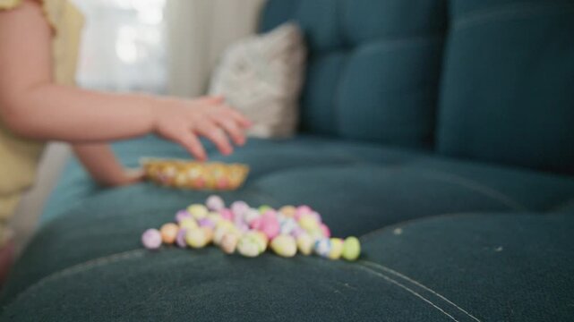 Child's hand pushes a basket of colorful candies across a couch. The basket tips, spilling candies onto the fabric. The child's hand moves gently, showing care and curiosity