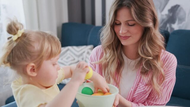 Mother helps her child eat breakfast together. Little girl smiles while holding spoon. Mother looks lovingly at her child. They share meal on cozy sofa. Perfect for family, parenting, morning routines