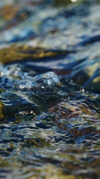 Close-up of fast-flowing, clear mountain stream water. Sunlight glints off rippling surface, revealing submerged stones. Water movement creates natural, dynamic textures. Ideal for nature, travel