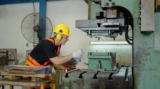 Industrial factory worker wearing safety helmet, glasses, and ear protection using spray bottle to clean and maintain heavy metal stamping press machine during industrial manufacturing process.