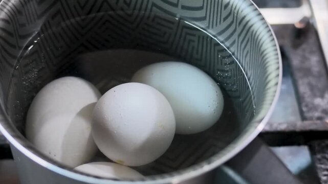 Static close-up shot of four white eggs resting inside a stainless steel cooking pan as water begins to boil on a domestic kitchen stove.