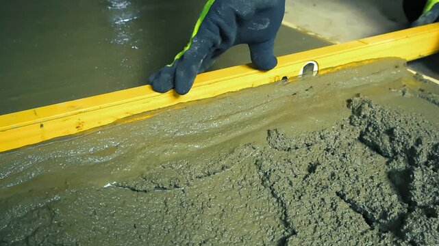Male builder using a spirit level to screed and flatten freshly poured wet cement mix on a floor, creating a smooth surface for a new screed during a house renovation or construction project