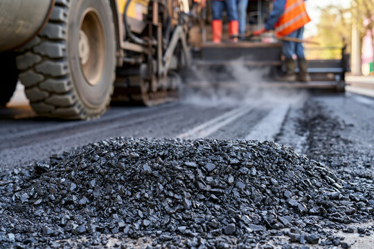 Heavy machinery compacts fresh asphalt. Workers in orange vests stand nearby. Black gravel piles beside wet pavement. Smoke rises from the machine's operation