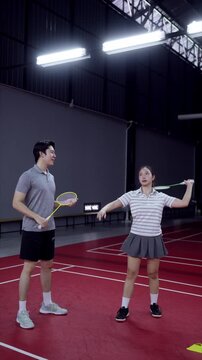 Asian male badminton coach guiding young woman through racket swing technique on indoor court
