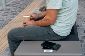 Fototapeta premium Man sitting on modern smart bench in city, holding coffee cup while his smartphone recharges on built-in wireless charging pad. Innovative urban infrastructure, street furniture, smart city lifestyle