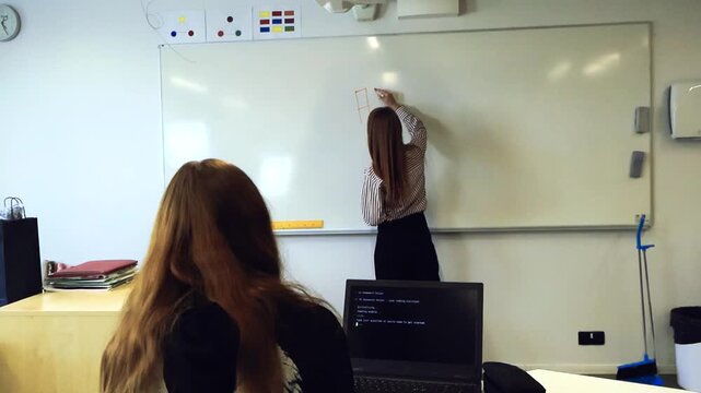 Female teacher explaining artificial intelligence concepts on a whiteboard while a student with a laptop follows the computer science lesson, focusing on machine learning and programming