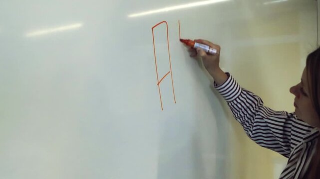 Young female teacher writing the words AI CLASS on a whiteboard with a red marker during a lesson at school, introducing the topic of artificial intelligence and modern technology education