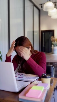 Stressed young Asian businesswoman holding head overwhelmed by paperwork at modern corporate office desk
