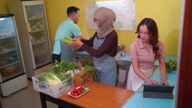 Three adults sort fresh vegetables in small neighborhood produce shop, woman in headscarf passes carrots to man near refrigerator shelves while another checks tablet beside crates.