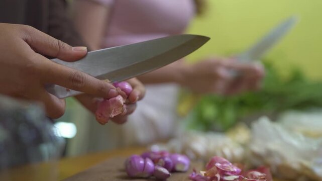 Close kitchen view shows hands trimming purple shallots with large knife over cutting board under warm indoor light, with wood surface and softly blurred cook in background near green wall.