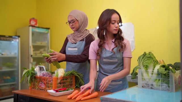 Two women in aprons organize fresh vegetables in small neighborhood produce shop, sorting red peppers, carrots, and leafy greens among baskets and crates under warm indoor lighting.