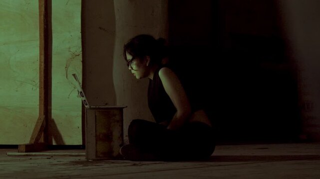 Wide profile shot of a female hacker or runaway sitting cross-legged on a dirty concrete floor in an abandoned, dark warehouse, working intently on her laptop.