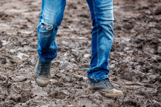 Person wears muddy jeans and boots. Feet step through wet, brown earth. Dirt clings to shoes and denim.