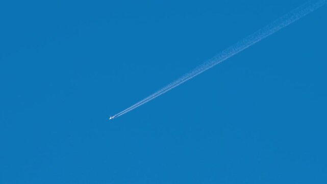 Passenger jet flying at cruising altitude leaves long white contrail across clear blue sky, sunny day, telephoto tracking shot