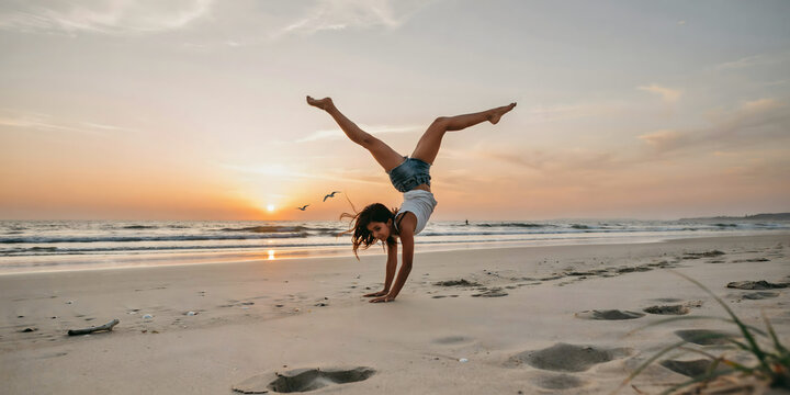 A young woman performing a handstand with legs in a split on the sandy beach during a golden sunset with the ocean in background