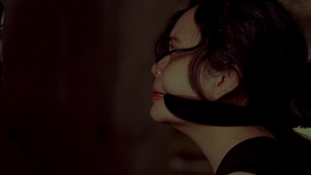 A terrifying moment where a gagged female prisoner forces a distressed, tearful smile while looking at her unseen kidnapper in a dark room.