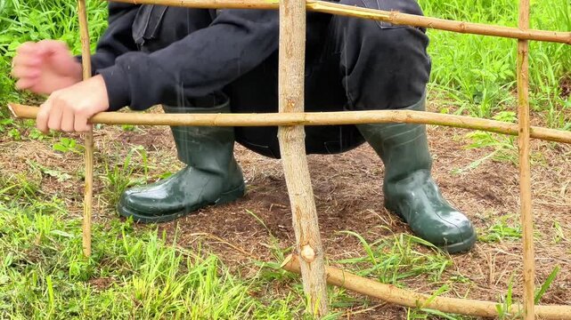 Person crouching in rubber boots and holding wooden branches to weave a wattle fence. Ideal for gardening guides, eco-friendly farming, DIY landscaping, and rural lifestyle media.