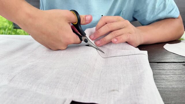 Close-up view of hands using scissors to cut marked light fabric on a table. Ideal for sewing tutorials, crafting media, textile design, and DIY project guides.