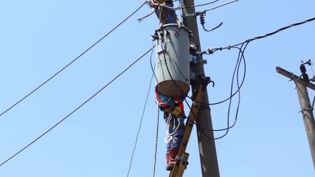 Electrician works high on a pole under clear sky. He secures transformer with tools and wires. Sunlight brightens his safety gear and tools. Worker focuses on task amid overhead lines