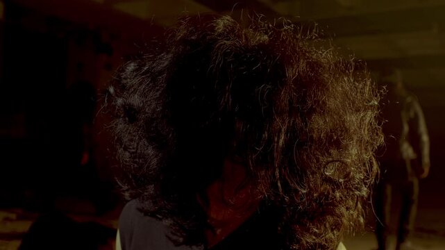 A moody close-up of an abducted woman's face hidden behind thick curly hair. She slowly raises her head with a traumatized look, while a blurry thug stands in the background.