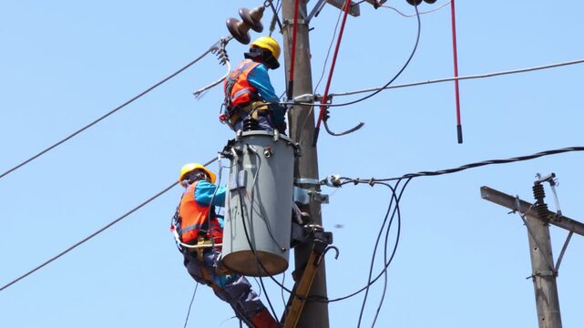 Electricians work on a tall pole. They wear hard hats and safety vests