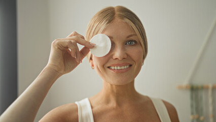 Woman holding cotton pad over eye in studio, smiling with exposed neckline and hand raised in skincare gesture  self care calm. © Krakenimages.com