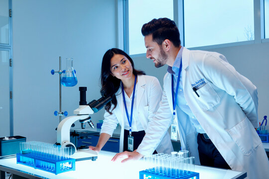 Asian young adult woman and Caucasian young adult man discussing research beside microscope and test tubes in laboratory. Ideal for healthcare innovation, teamwork, biotech marketing