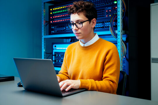 Young adult Caucasian woman working on laptop beside server racks, monitoring network systems and managing IT operations. Ideal for cybersecurity, data center, cloud computing marketing