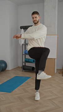 Man balancing on one leg on blue yoga mat while raising arms in a fitness studio with shelf and whiteboard; balance focus.
