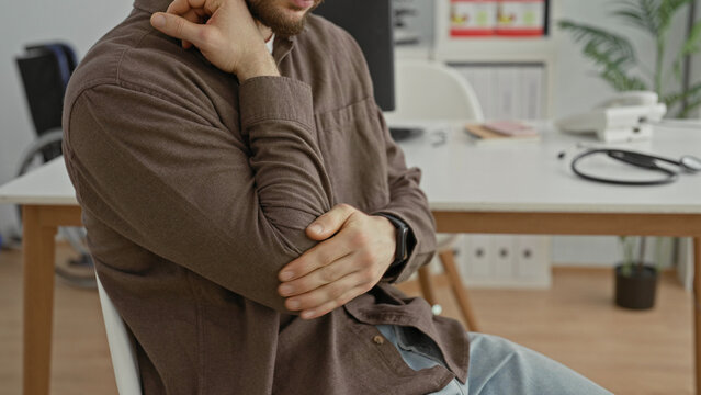 Man holding elbow and touching neck beside stethoscope on desk in medical building; discomfort seeking help.