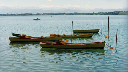 Rowing wooden boats moored to wooden poles with orange buoys on tranquil turquoise lake water against distant hills and mountains on a sunny day. © Ekaterina