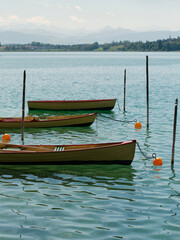Rowing wooden boats moored with chains to poles and orange buoys floating on clear turquoise lake water against distant hills and mountains on a sunny day. © Ekaterina