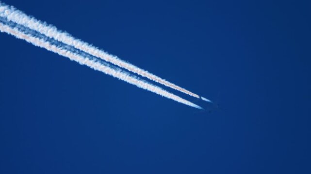 Passenger jet at cruising altitude leaves two parallel white contrails across deep blue sky, clear day, telephoto tracking shot