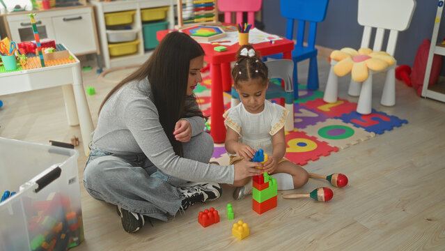 Woman stacking colorful blocks with little girl on foam playmat inside a building; playful learning.