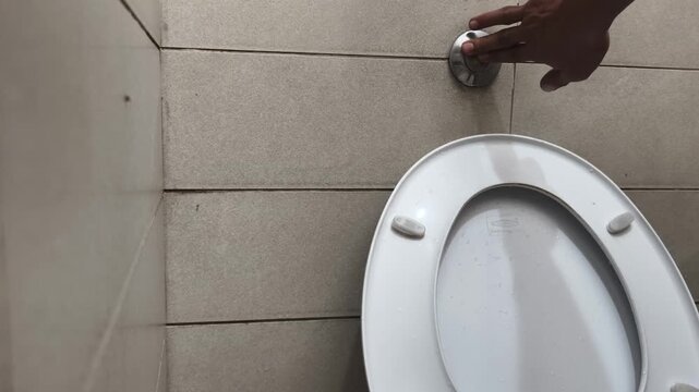 A close-up shot of a hand pressing a circular, wall-mounted flush button above a white ceramic toilet bowl in a modern bathroom with tiled walls, but the toilet fails to flush water.
