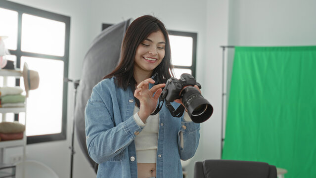 Young woman photographer adjusts a dslr camera and smiles while reviewing shots in a studio with green screen and softbox lighting; creative joy.