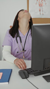 Woman in lavender medical scrubs with stethoscope at computer tilting head back and stretching neck at desk in building; fatigue stress recovery.