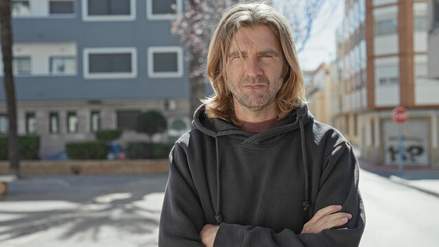 Man with long blond hair and arms crossed, smiling with hands visible, wearing a dark hoodie while standing on a sunny urban street in front of buildings; everyday contentment.