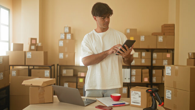 Man holding tablet and typing on laptop at a packing station surrounded by stacked parcels, barcode scanner and red coffee cup in a building; focused productivity.