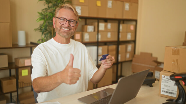 Man holding creditcard and typing on laptop while packing parcels at a small shipping building with barcode scanner, stacked boxes and desk supplies; entrepreneurial confidence.