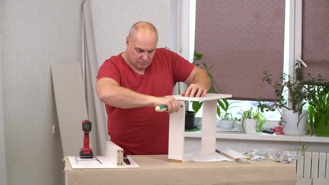 A man focuses on assembling a piece of furniture in a bright room filled with plants. He uses a drill and various tools while carefully measuring and securing pieces together.