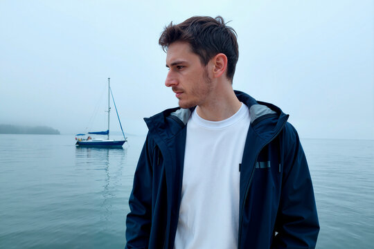 Young adult Caucasian man standing by calm lake, looking away near sailboat and reflecting on travel freedom and solitude. Usable for tourism, lifestyle, mindfulness marketing