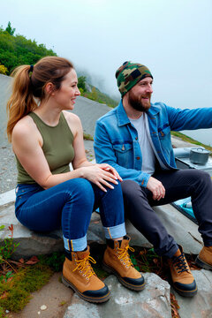 Young adult Caucasian couple sitting outdoors, smiling and pointing toward mountain view during hiking break. Ideal for travel marketing, adventure lifestyle, weekend getaway campaigns