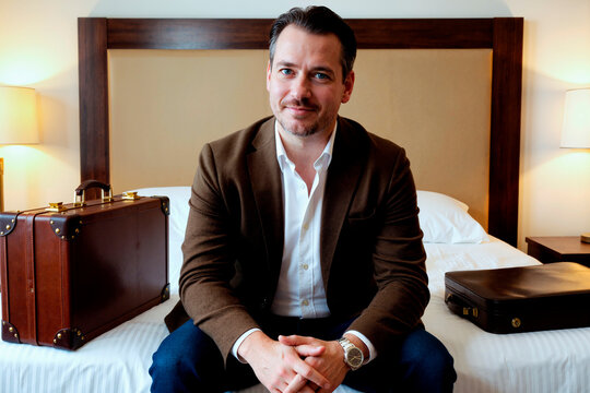 Portrait of middle aged Caucasian businessman sitting on hotel bed beside luggage, preparing for business travel or work trip. Useful for corporate travel, mobility, hospitality marketing
